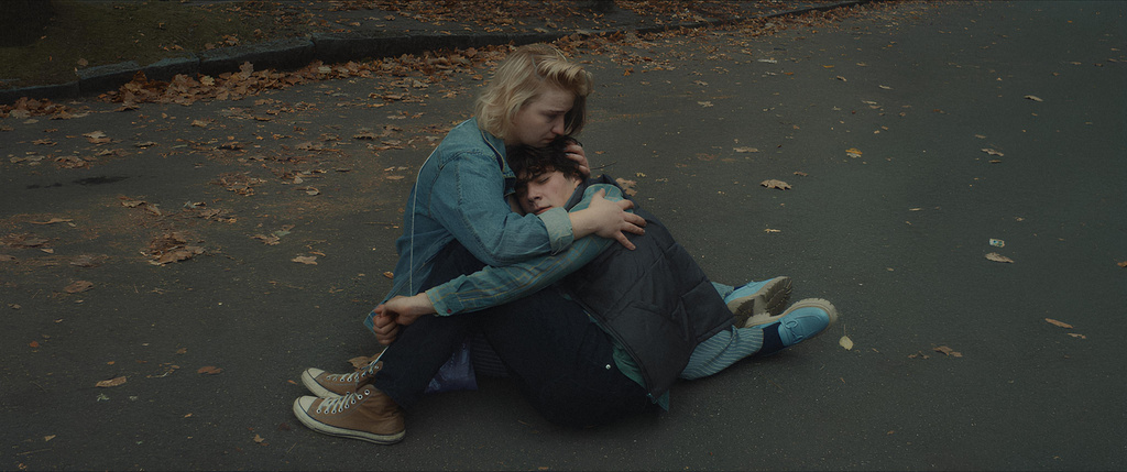 Filmszene: zwei Menschen sitzen eng beieinander am Boden zwischen herbstlichem Laub