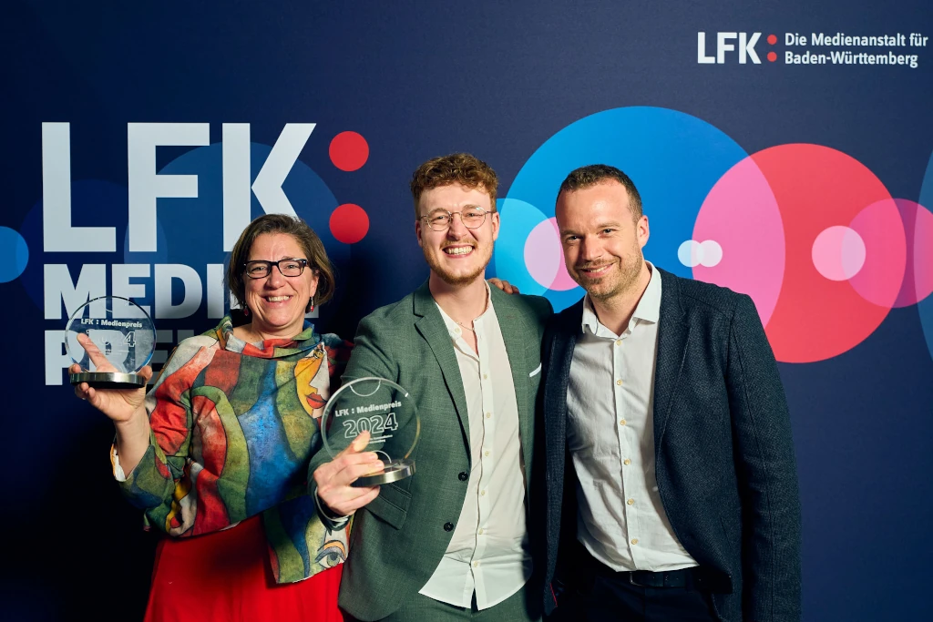 Preisverleihung des LFK-Medienpreises 2024: drei Personen mit Glastrophäen vor einer Wand mit dem Logo der Landesanstalt für Kommunikation Baden-Württemberg