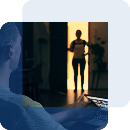 Ein Mann sitzt im abgedunkelten Zimmer vor einem Laptop, im hellen Türrahmen dahinter steht eine zweite Person